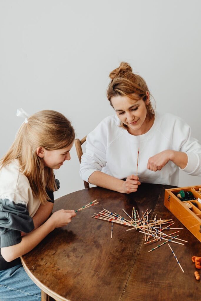 A cheerful scene of a mother and daughter playing a stick game at a wooden table indoors.